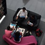 Grupo diverso de adultos colaborando com laptops em uma área de convivência de uma biblioteca.